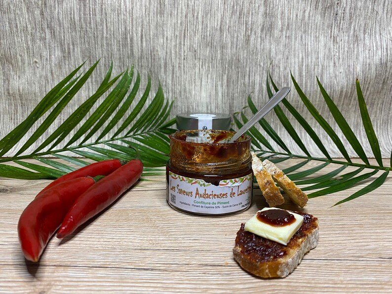 Confiture de piment dans un pot ouvert avec cuillère, entourée de piments rouges et toast léger au pain, pour comment faire de la confiture de piment.